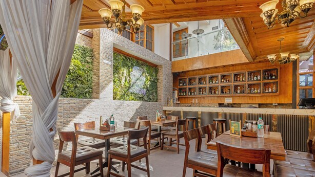 Un restaurante con mesas y sillas de madera, una zona de bar con estantes llenos de botellas y un amplio ventanal con plantas.