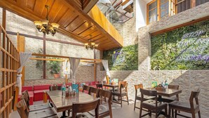 Un restaurante con mesas y sillas de madera, una zona de asientos con asientos tipo banca tapizados en rojo, y una pared con plantas y un letrero de neón.