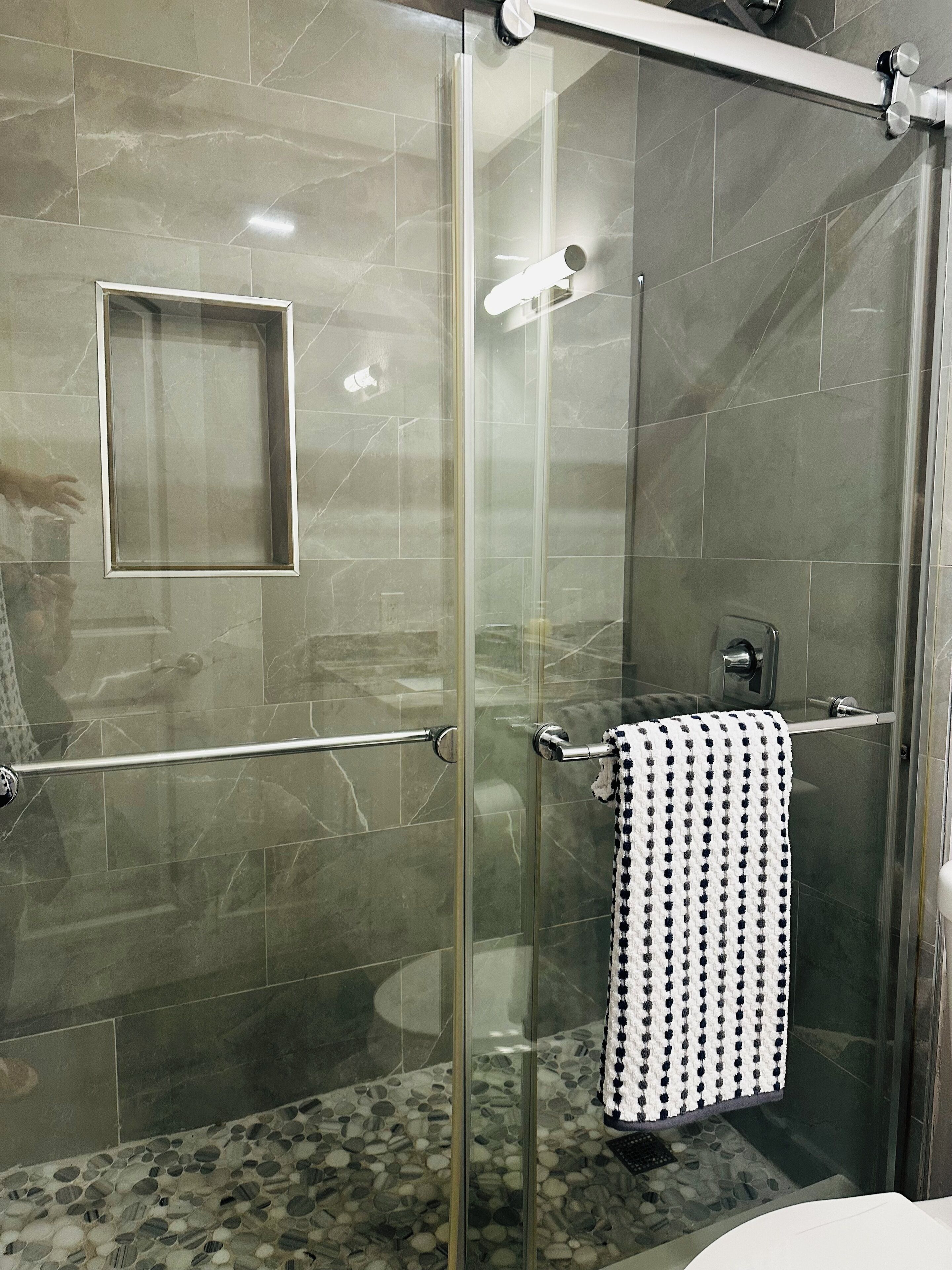 Une douche fermée par des parois de verre, une barre à serviettes en métal, un porte-serviettes avec une serviette à motifs et un petit miroir carré fixé au mur.