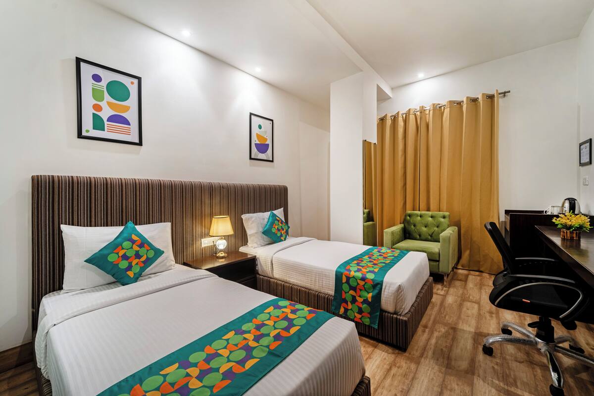 A hotel room with two beds, a green armchair, a desk with a chair, and a curtain.