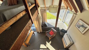 A loft-style bedroom with a bunk bed, a small sofa, a chair, and a staircase leading to an outdoor area. - Chalet 6 - Ohakune Holiday Home (Ohakune)