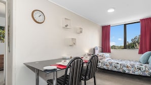 Une pièce de taille modeste comprenant une table à manger, des chaises, un lit recouvert d’une couvre-lit à motifs floraux, une horloge et trois étagères murales ornées d’objets décoratifs.