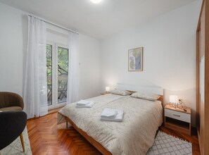 Cozy retreat with a large bed, nightstand, chair, and table, set against a clean, minimalist backdrop.
