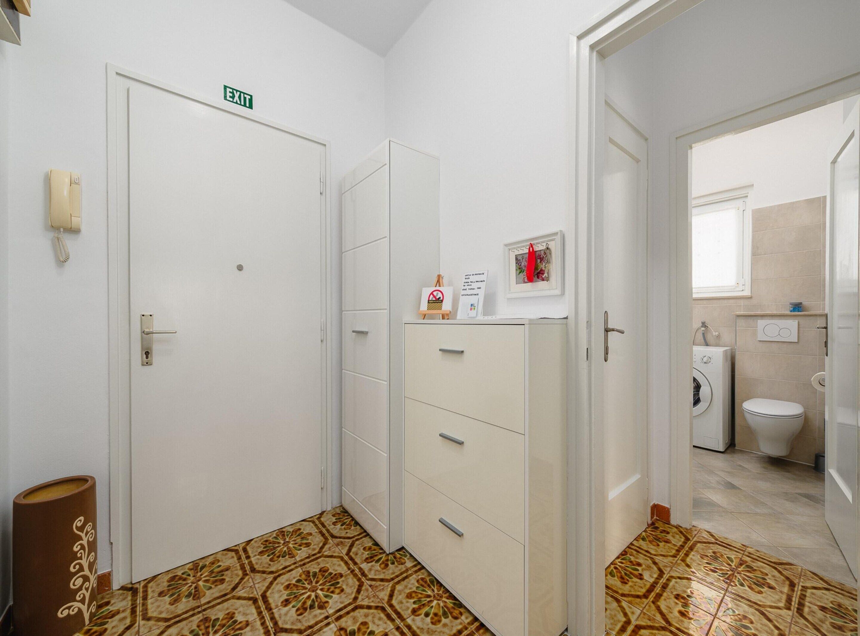 A hotel room with a white door marked "EXIT," a small cabinet with drawers, a telephone, and a trash bin.