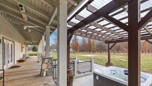 Ein überdachter Außenbereich mit Holzterrasse, Whirlpool, Steinpfeilern und Blick auf einen Park mit Bäumen.