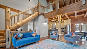 Ein geräumiger Wohnbereich mit blauem Sofa und Sesseln, einer Holztreppe und einer Küche im Hintergrund.
