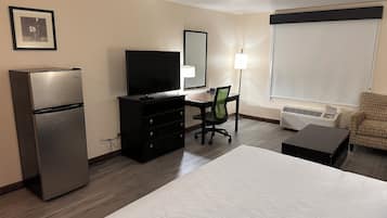 A hotel room with a bed, a desk with a chair, a television, a refrigerator, a lamp, and a window with blinds.