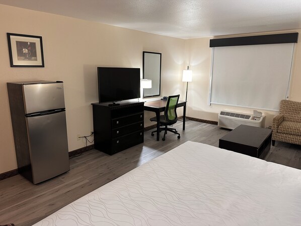 Habitación de hotel con cama, escritorio con silla, televisión, refrigerador, lámpara y ventana con persianas.