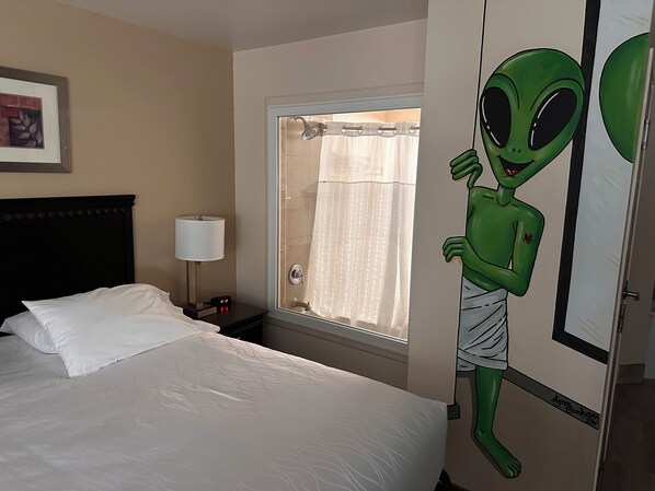 Una habitación de hotel con cama, mesita de noche, lámpara, ventana con cortina de ducha e ilustración de un extraterrestre verde en la pared.