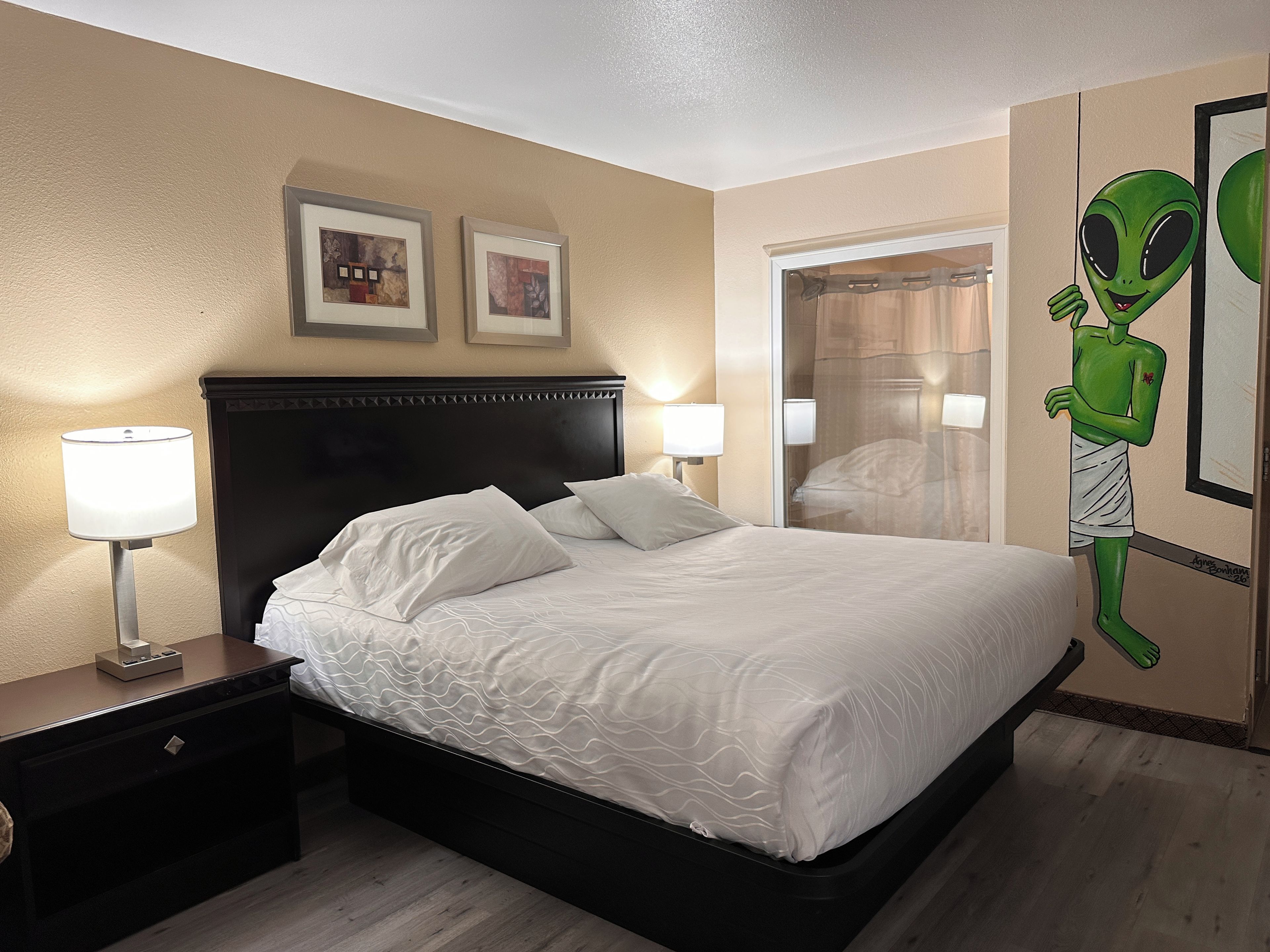 Une chambre d’hôtel avec un lit, deux lampes de chevet, une table de nuit, un miroir et un objet de décoration représentant un extraterrestre vert.
