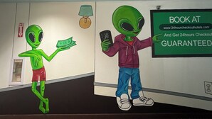 A hallway with alien characters, a fire exit sign, a lamp, and a sign advertising a 24-hour checkout guarantee. - CASH REWARDS INN & SUITES (Roswell)
