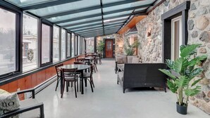 A covered outdoor seating area with tables and chairs, stone walls, and a view through large windows.