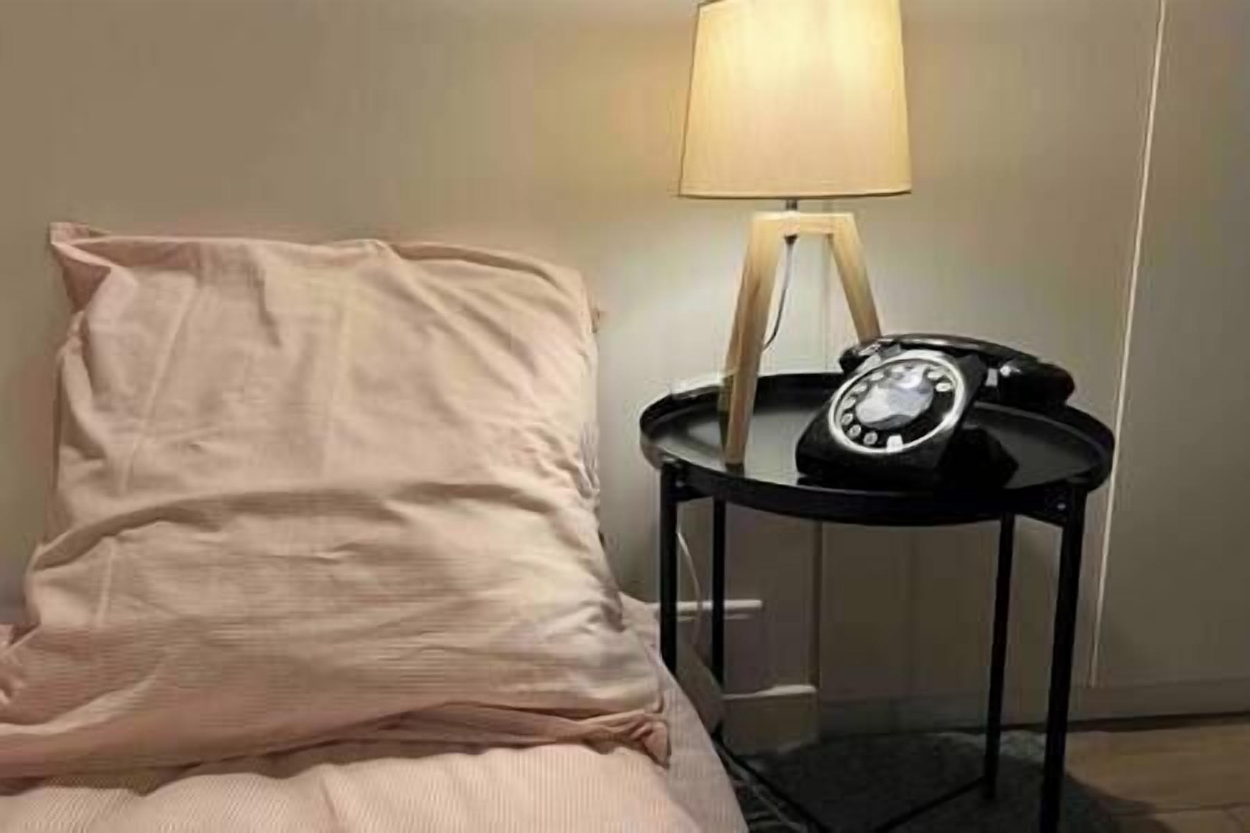 A bed with a white pillow and a black bedside table with a lamp and a rotary phone.