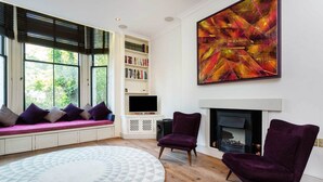 Een moderne woonkamer met een open haard, een groot abstract schilderij, een inbouwboekenplank en een gezellige zithoek met een kamerkleed met dessin.