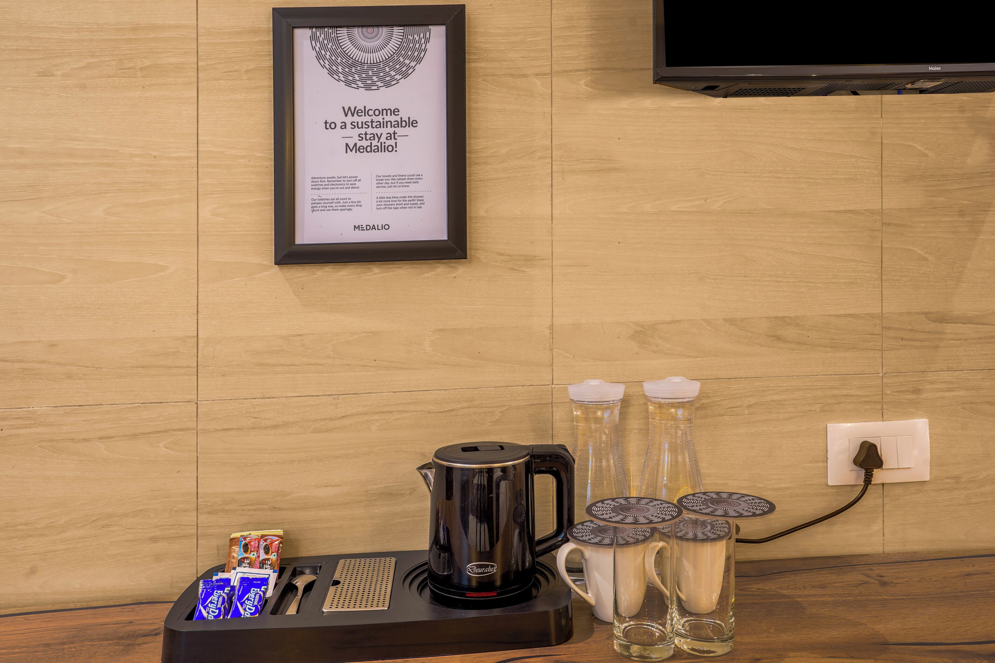 A framed welcome sign, a coffee maker, a mug, and a bottle of water on a wooden table.