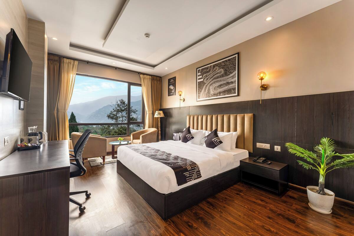 Quarto de hotel moderno com uma cama grande, uma mesa, uma cadeira, uma planta em vaso e vista para as montanhas.