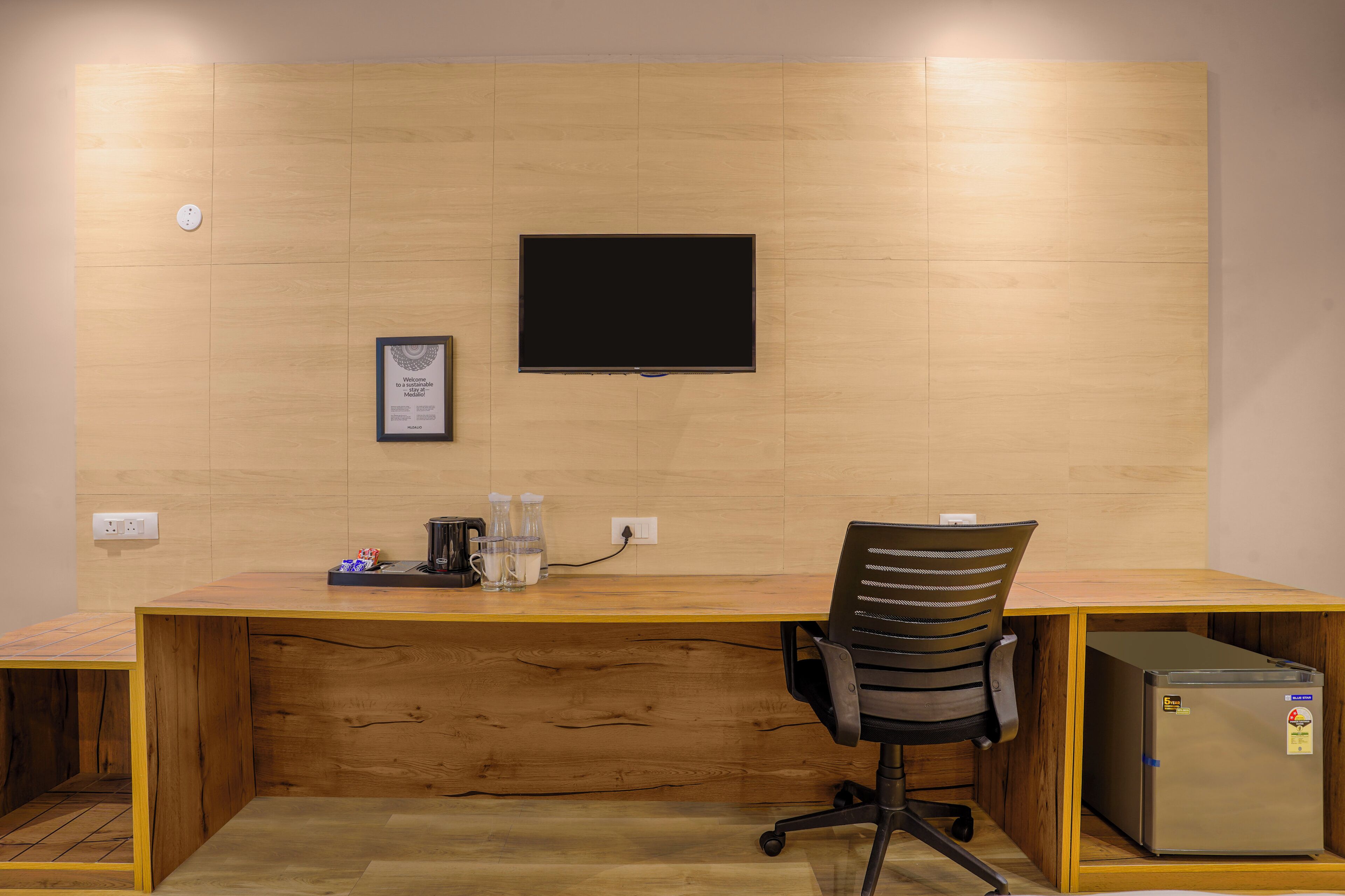 A wooden desk with a computer, a chair, a small refrigerator, and a flat-screen TV mounted on the wall.