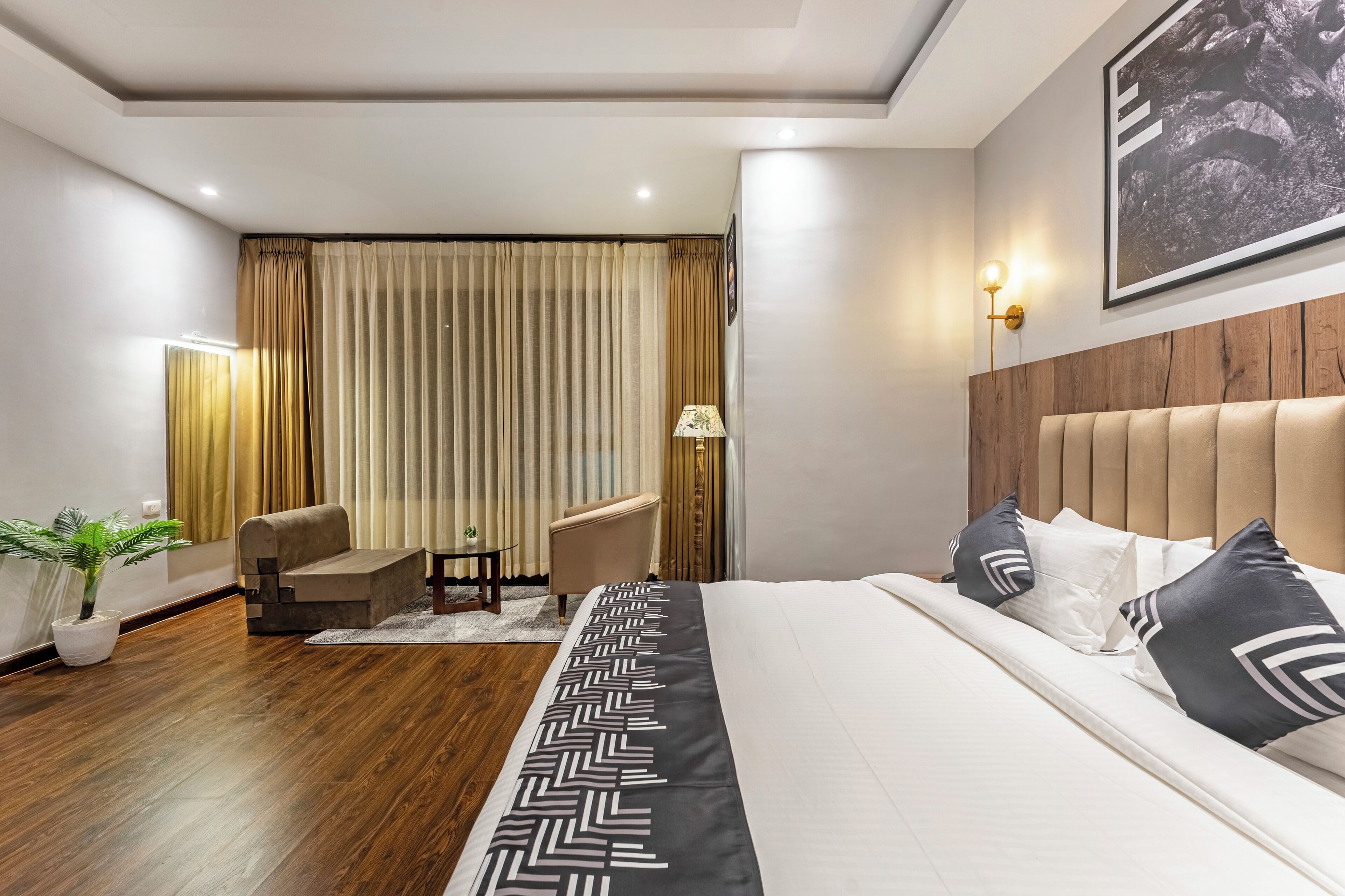 Une chambre d’hôtel avec un grand lit, une tête de lit, un coin salon avec une chaise et une table, une plante en pot et un rideau.