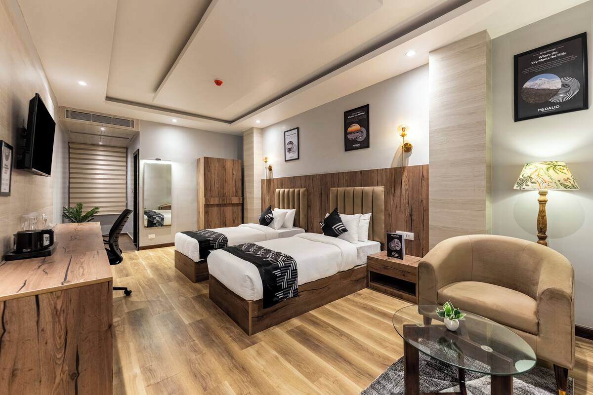 Quarto de hotel moderno com piso de madeira, duas camas com cabeceiras, uma mesa de escritório com cafeteira, um sofá e uma mesa de vidro com uma planta.