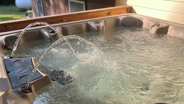 A hot tub with water flowing into it, surrounded by a wooden deck and a house in the background.