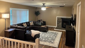A living room with a sectional sofa, a fireplace, a ceiling fan, and a large window with blinds.