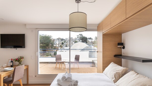 Una camera d'albergo con un letto, una scrivania con una sedia, una TV a parete e un balcone con sedie e un tavolo.