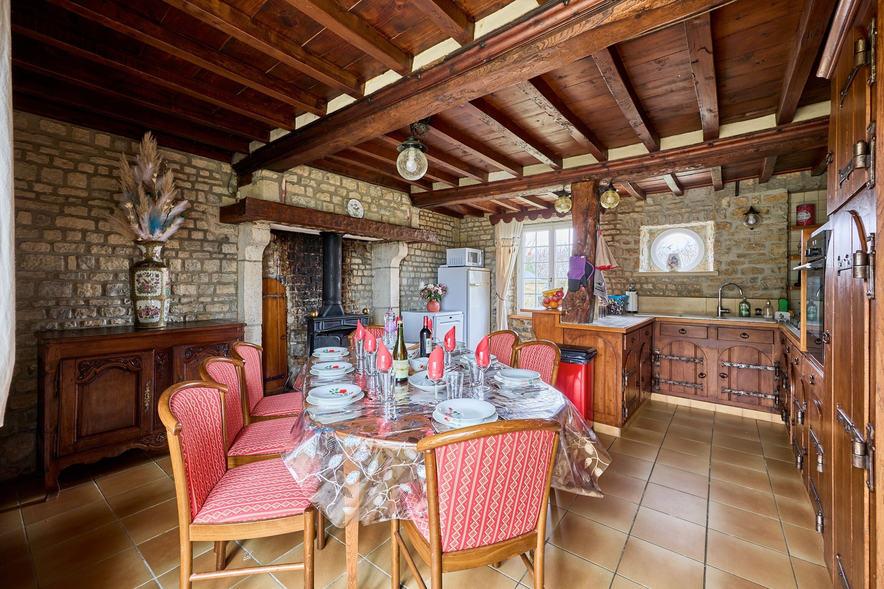 Un comedor con una mesa redonda dispuesta para cuatro personas, rodeada de sillas de madera con tapicería estampada. Se aprecia una pared de piedra y vigas de madera al fondo.
