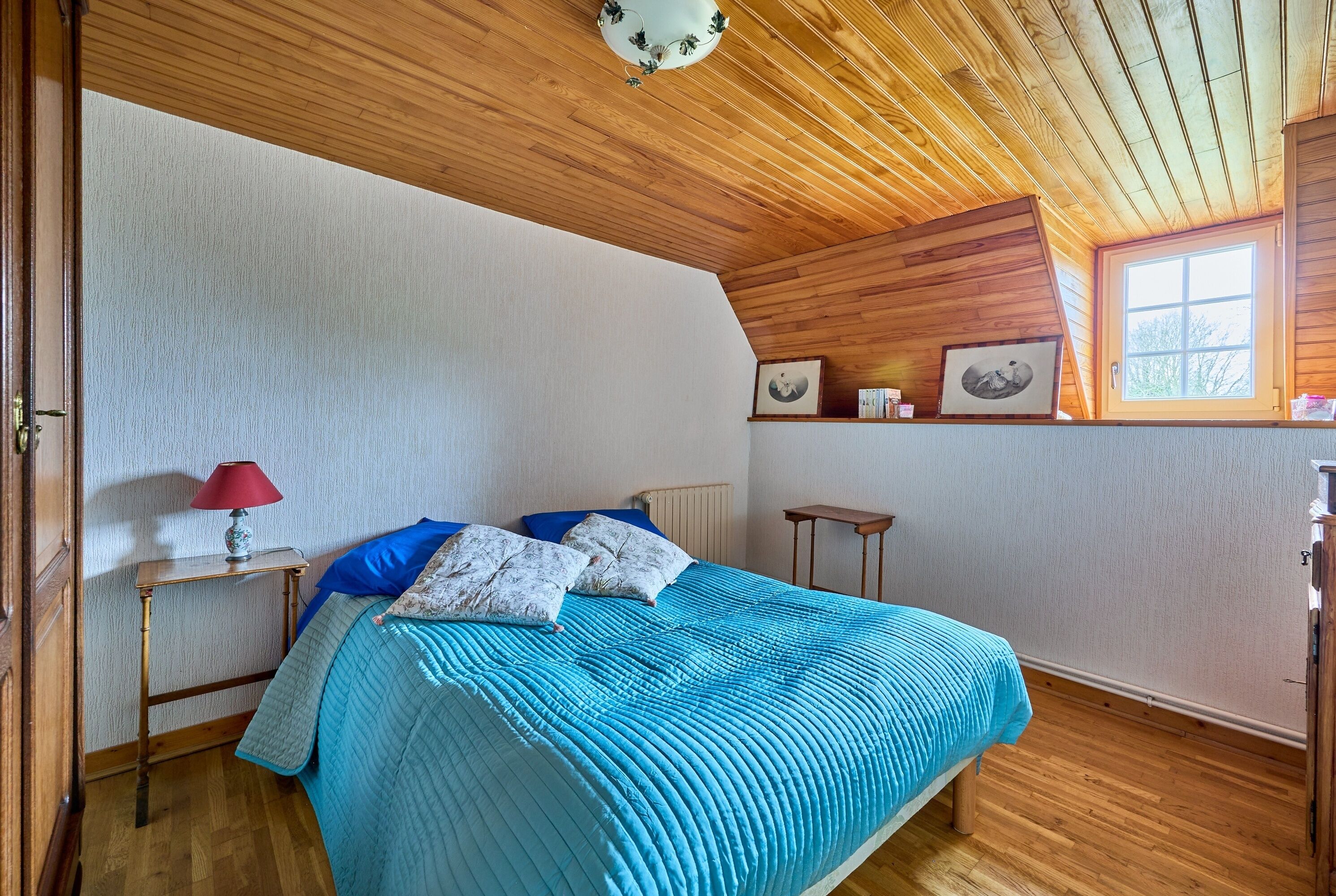 A bedroom with a wooden ceiling, a bed with a blue quilt, a red lamp, and framed pictures on the wall.