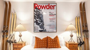 Une chambre d’hôtel avec une grande affiche du magazine « Powder », des paires de skis accrochés au mur, un lit avec une literie blanche et des oreillers orange, et deux tables de chevet avec des lampes.