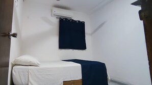 Une petite chambre avec un lit simple, une couvre-lit blanche, un rideau bleu foncé et un appareil de climatisation fixé au mur.