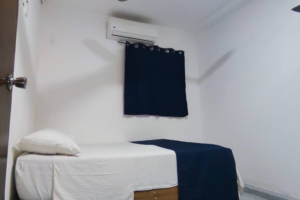 A small, single-bed room with a white bedspread, a dark blue curtain, and an air conditioning unit on the wall.