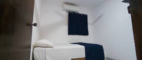 A small, single-bed room with a white bedspread, a dark blue curtain, and an air conditioning unit on the wall.