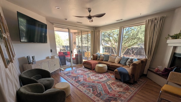 A spacious living room with a large sofa, a coffee table, and a patterned rug. There is a television mounted on the wall, and the room has large windows with curtains.