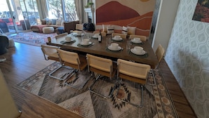 A dining area with a large table set for six, surrounded by wicker chairs, with a view of the outdoors through large windows.