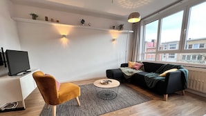 A modern living room with a sofa, a chair, a coffee table, a TV, and a large window with a view of buildings. - Bright & Stylish in Nature (Brussels)