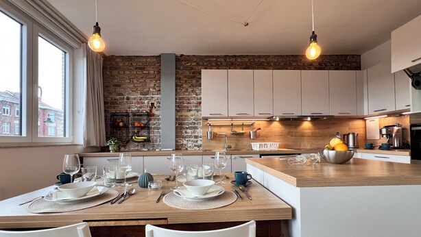 A modern kitchen with a brick wall, wooden countertops, and white cabinets. There is a dining area with a table set for two, and a window providing natural light. - Bright & Stylish in Nature (Brussels)