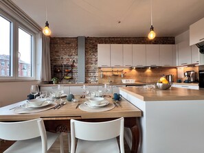 A modern kitchen with a brick wall, wooden countertops, and white cabinets. There is a dining area with a table set for two, and a window providing natural light. - Bright & Stylish in Nature (Brussels)