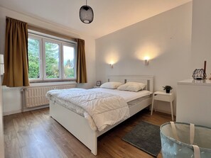 A bedroom with a double bed, a bedside table, a nightstand, a window with curtains, a radiator, a hanging light fixture, and a small plant on the nightstand. - Bright & Stylish in Nature (Brussels)