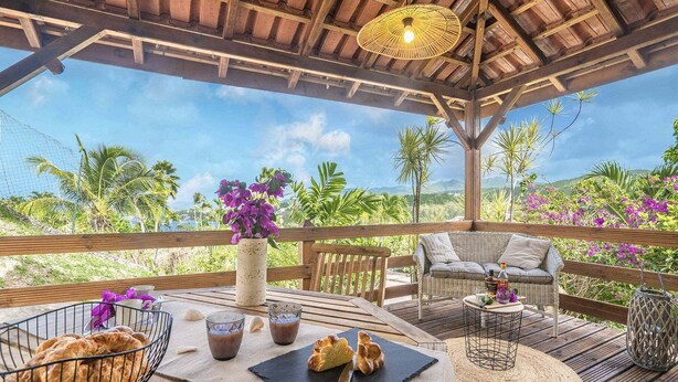 Un espace extérieur aménagé avec une terrasse en bois, un canapé en osier, une table garnie de nourriture et de boissons, et une vue sur les palmiers et les montagnes.