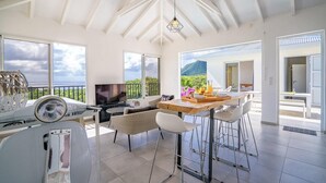 A spacious living area with a large TV, comfortable seating, a dining table with fruit, and a scenic mountain view. - Domaine Agapé - Four Bedroom Villa, Sleeps 9 (Le Diamant)