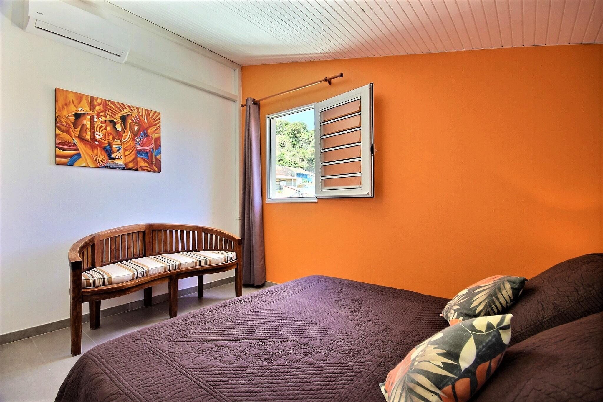 Une chambre avec un mur orange, un lit recouvert d’une couverture marron, un banc en bois et un tableau.