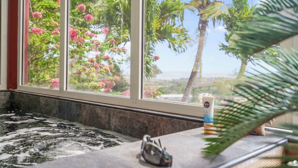 A bathroom with a bathtub, a window with a view of pink flowers and palm trees, and a bottle of lotion on the counter.