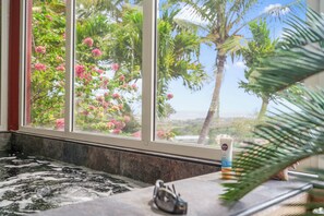 Ein Badezimmer mit Badewanne, einem Fenster mit Blick auf rosa Blüten und Palmen sowie einer Lotionflasche auf der Ablagefläche.
