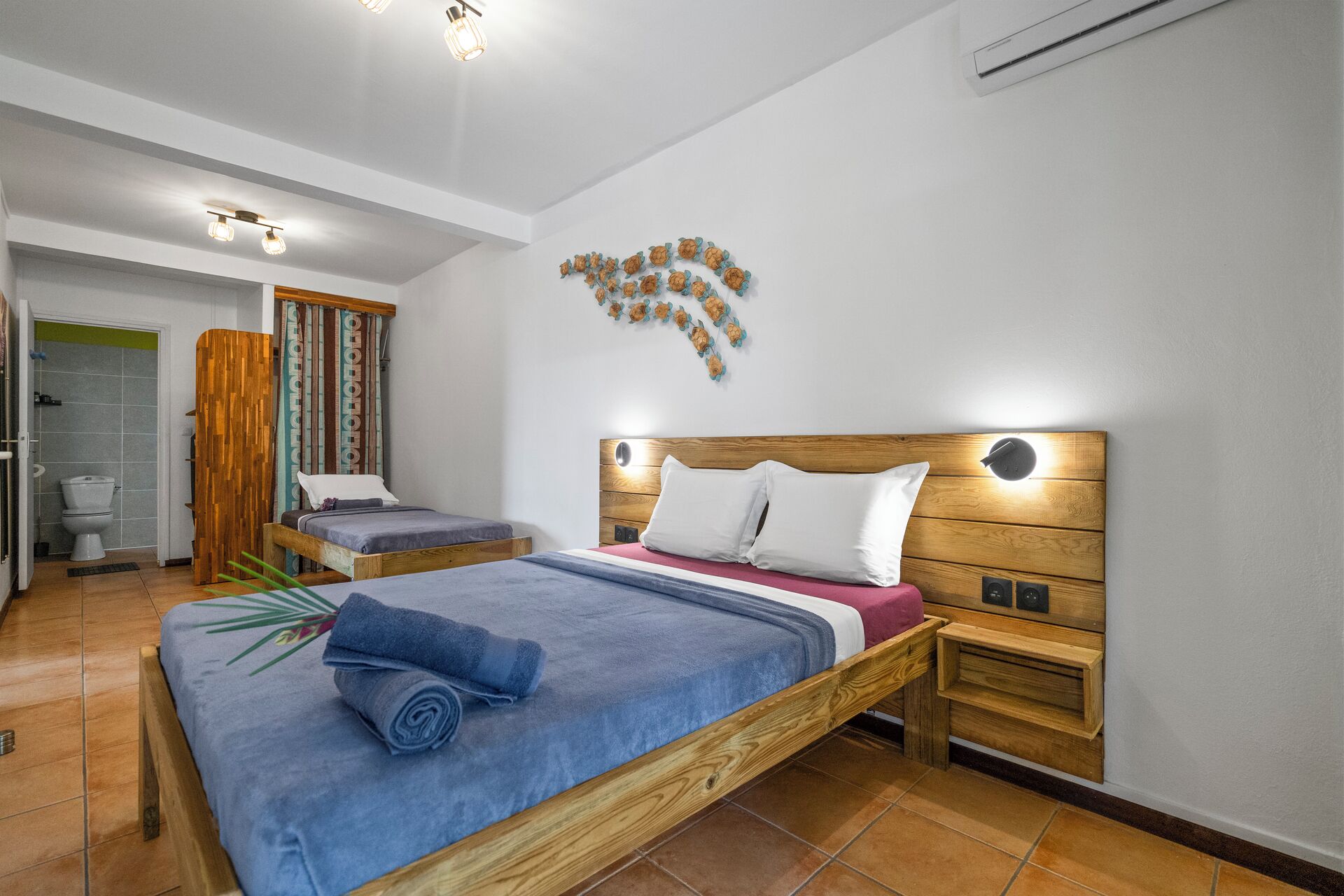 A bedroom with two beds, a wooden headboard, a wall-mounted light, and a bathroom visible through an open door.
