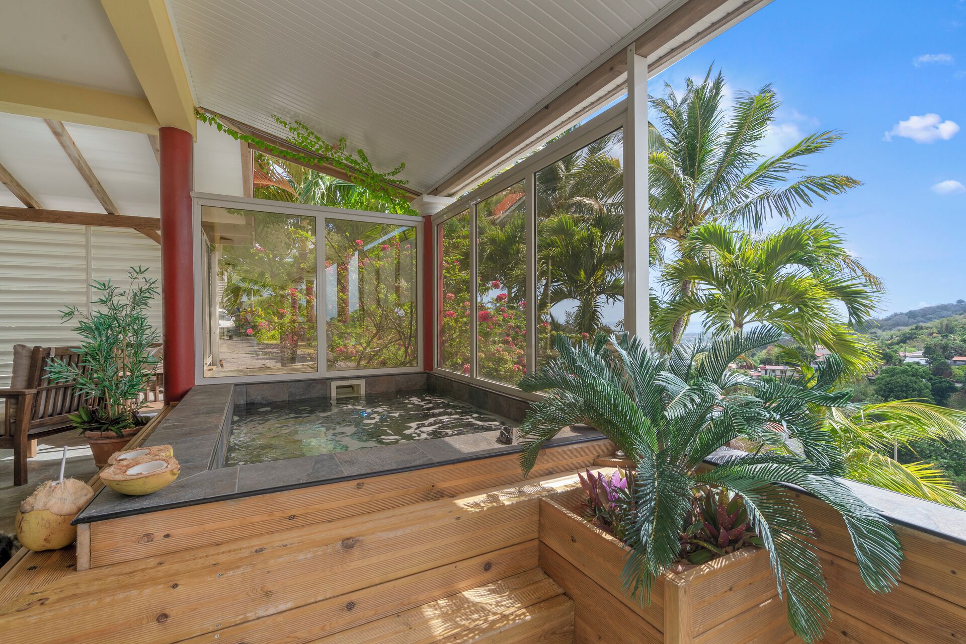 Une terrasse en bois dotée d’un jacuzzi, entourée de plantes en pot et de grandes fenêtres offrant une vue sur des palmiers et une colline.