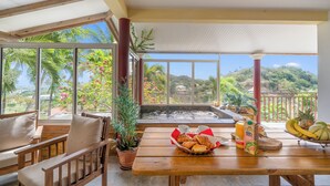 A wooden table with a basket of croissants, a bottle of juice, and a pineapple on a balcony with a view of lush greenery and hills.
