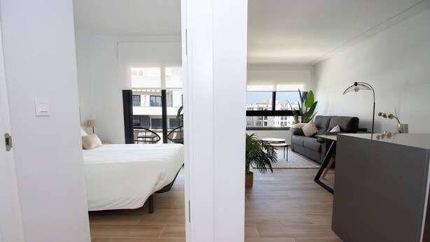 Un apartament modern amb llit, sofà, taula de dinar i un balcó amb plantes.