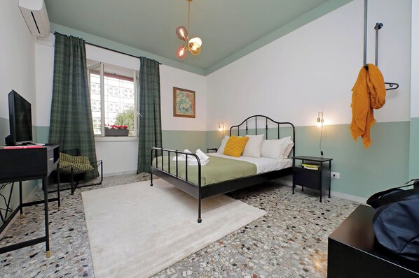 A bedroom with a bed, a desk, a chair, a window with curtains, a hanging towel, and a wall-mounted light fixture. - TM APARTMENT (Roma)