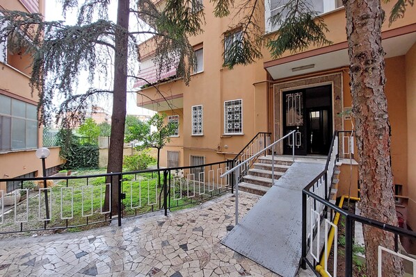A multi-story building with a staircase leading to the entrance, surrounded by trees and a fenced garden area. - TM APARTMENT (Roma)