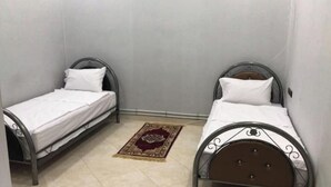 Deux lits simples avec des structures en métal et du linge de lit blanc, un petit tapis et des murs de couleur neutre.